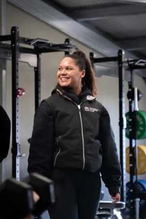 A smiling woman in a gym holds a dumbbell, showcasing her strength and enthusiasm for fitness.
