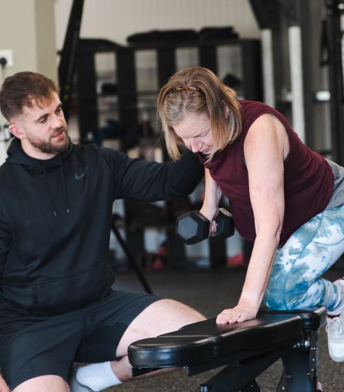 A woman exercises with a dumbbell on a bench, assisted by a trainer in a gym. The setting is supportive and focused, with gym equipment in the background.