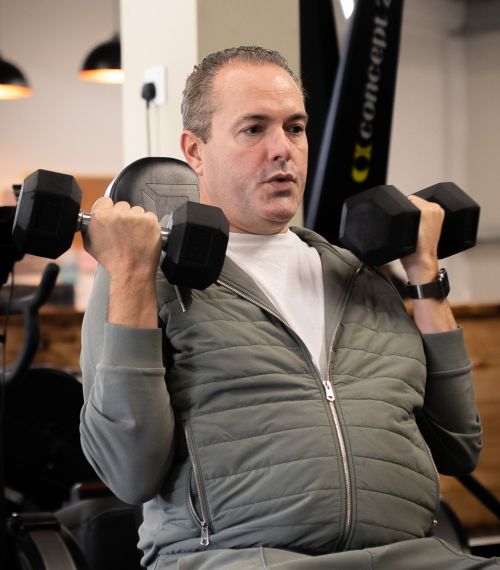 A man in a gym performs shoulder presses with dumbbells. He is wearing a gray tracksuit and appears focused and determined, conveying a sense of concentration.